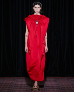 Model walking the runway in a bold red, voluminous cape-style dress with a statement necklace.