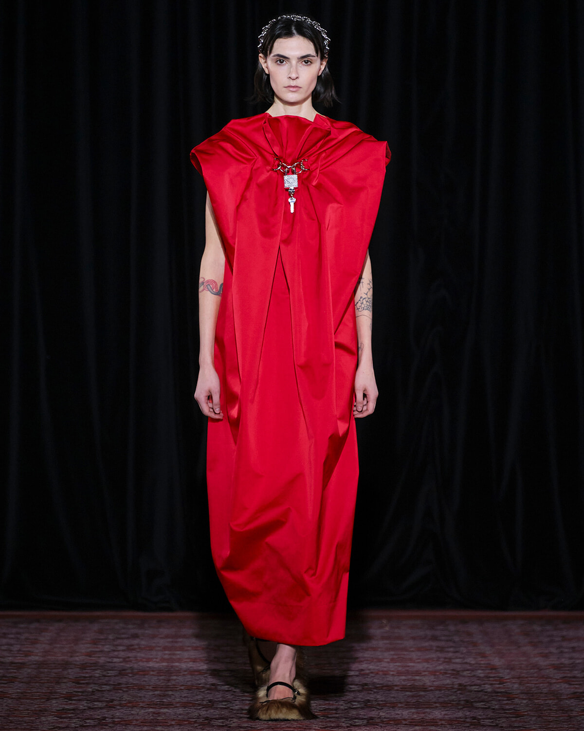 Model walking the runway in a bold red, voluminous cape-style dress with a statement necklace.