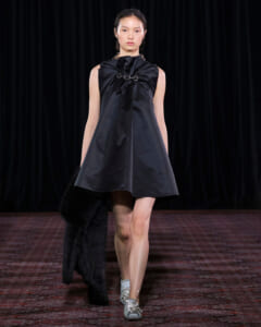 Model on a runway wearing a sleeveless dark gray A-line dress with a gathered neckline and chain detail, carrying a black fur coat.