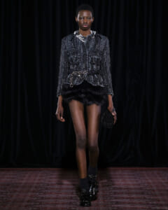 Runway model wearing a metallic gray tweed jacket with a black fur mini skirt and chunky belt, holding a beaded clutch.
