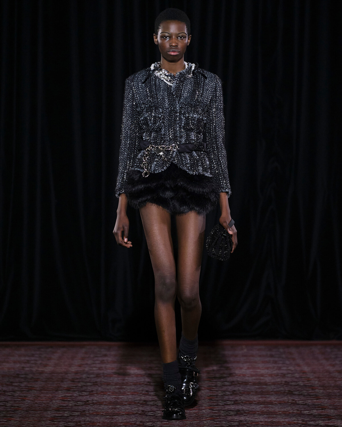 Runway model wearing a metallic gray tweed jacket with a black fur mini skirt and chunky belt, holding a beaded clutch.