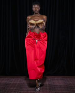 Model walks a runway wearing a brown fur bandeau and a voluminous red midi skirt with a metal belt accessory against a black backdrop.