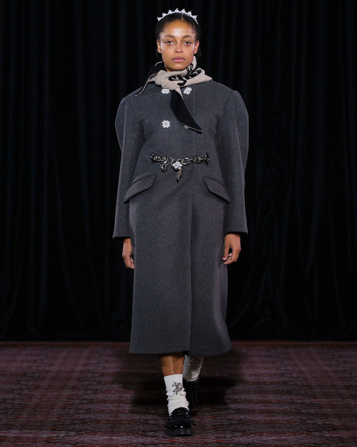 Model walking a runway in a dark gray wool coat with a jeweled belt and scarf, wearing a small tiara headpiece.