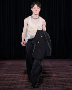 Model walking a fashion runway in a sheer white tank with floral appliqué, oversized black trousers, and a black blazer draped over the arm, accessorized with a chunky silver necklace and earrings.