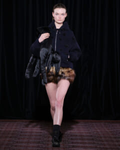 Model on a runway wearing a navy oversized hoodie with bead embellishments and brown fur shorts, holding long black gloves.