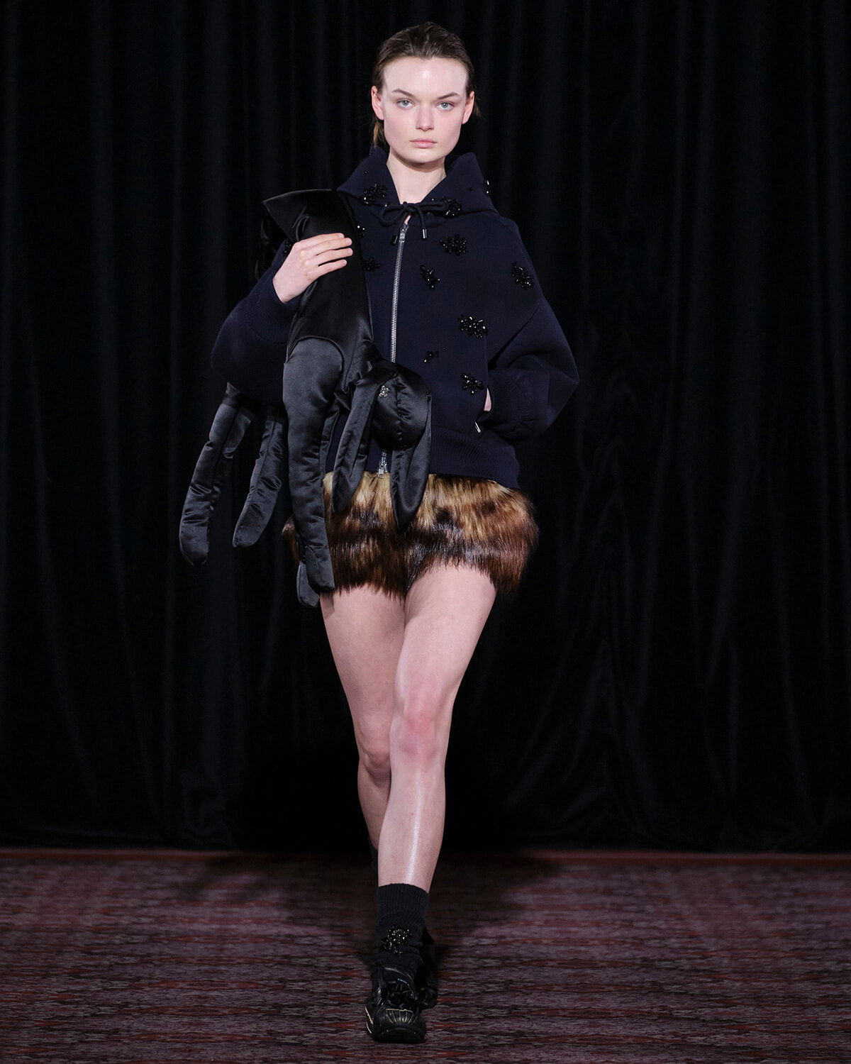 Model on a runway wearing a navy oversized hoodie with bead embellishments and brown fur shorts, holding long black gloves.