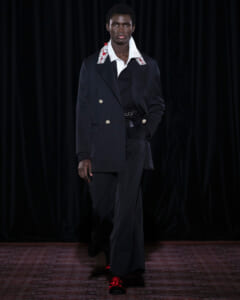 Male model walking a runway in a navy double-breasted suit, white embellished collar, and matching trousers with red-accent shoes.