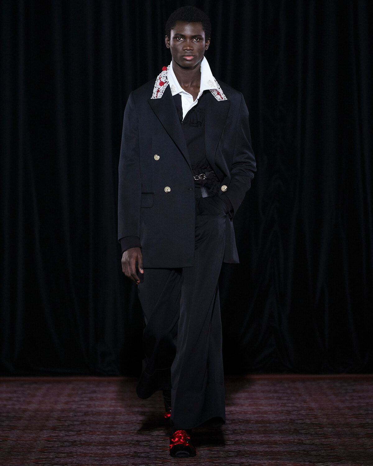 Male model walking a runway in a navy double-breasted suit, white embellished collar, and matching trousers with red-accent shoes.
