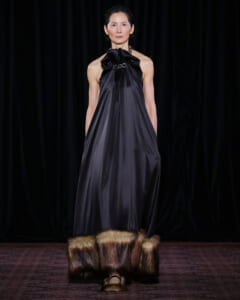 Model on a runway wearing a dark satin halter gown with a large bow at the neckline and fur-trimmed hem, black backdrop behind.