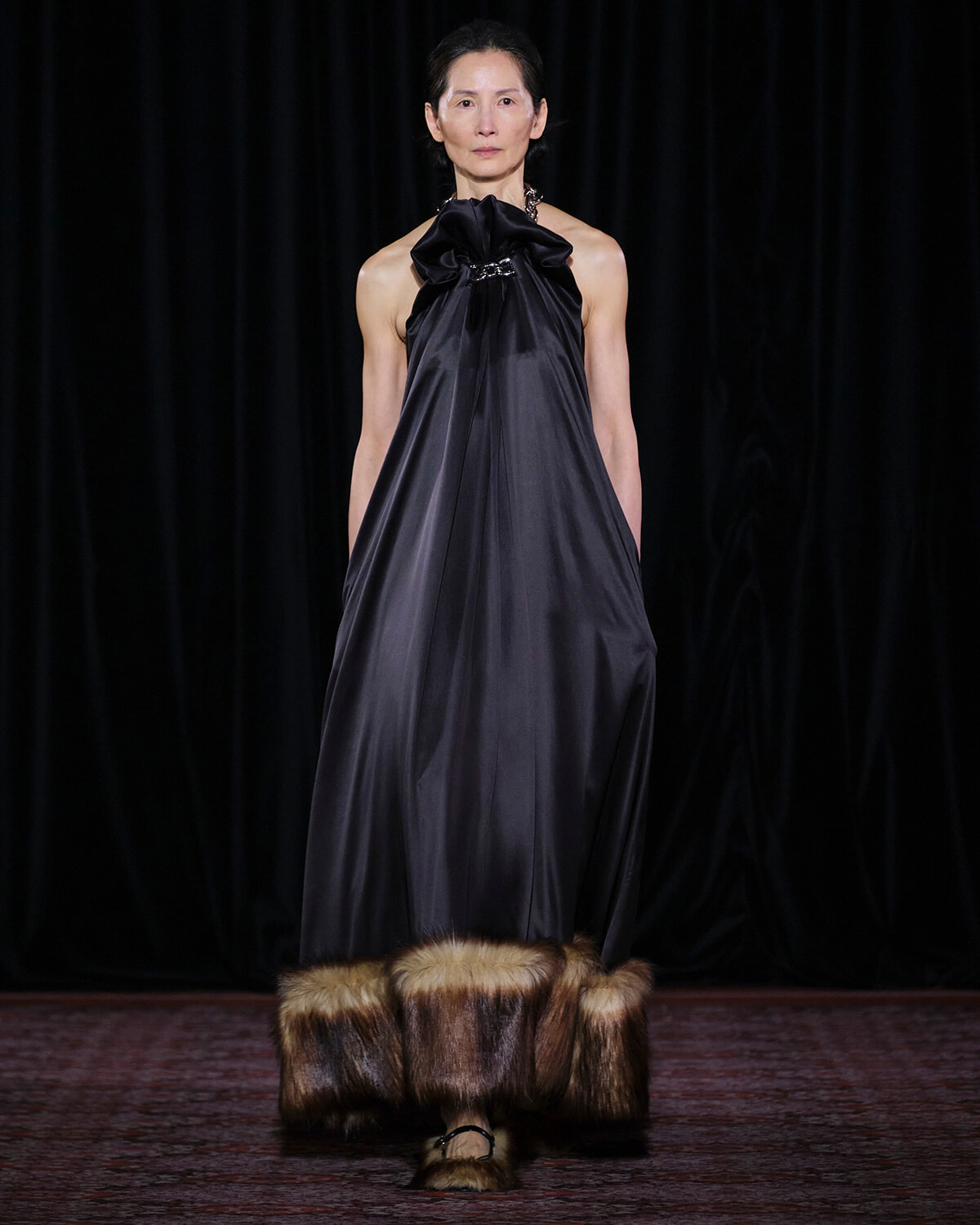 Model on a runway wearing a dark satin halter gown with a large bow at the neckline and fur-trimmed hem, black backdrop behind.