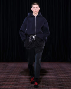 Male model walks the runway in an oversized navy hoodie with front zipper, black pants, a silver chain belt, and black shoes with red floral decorations.
