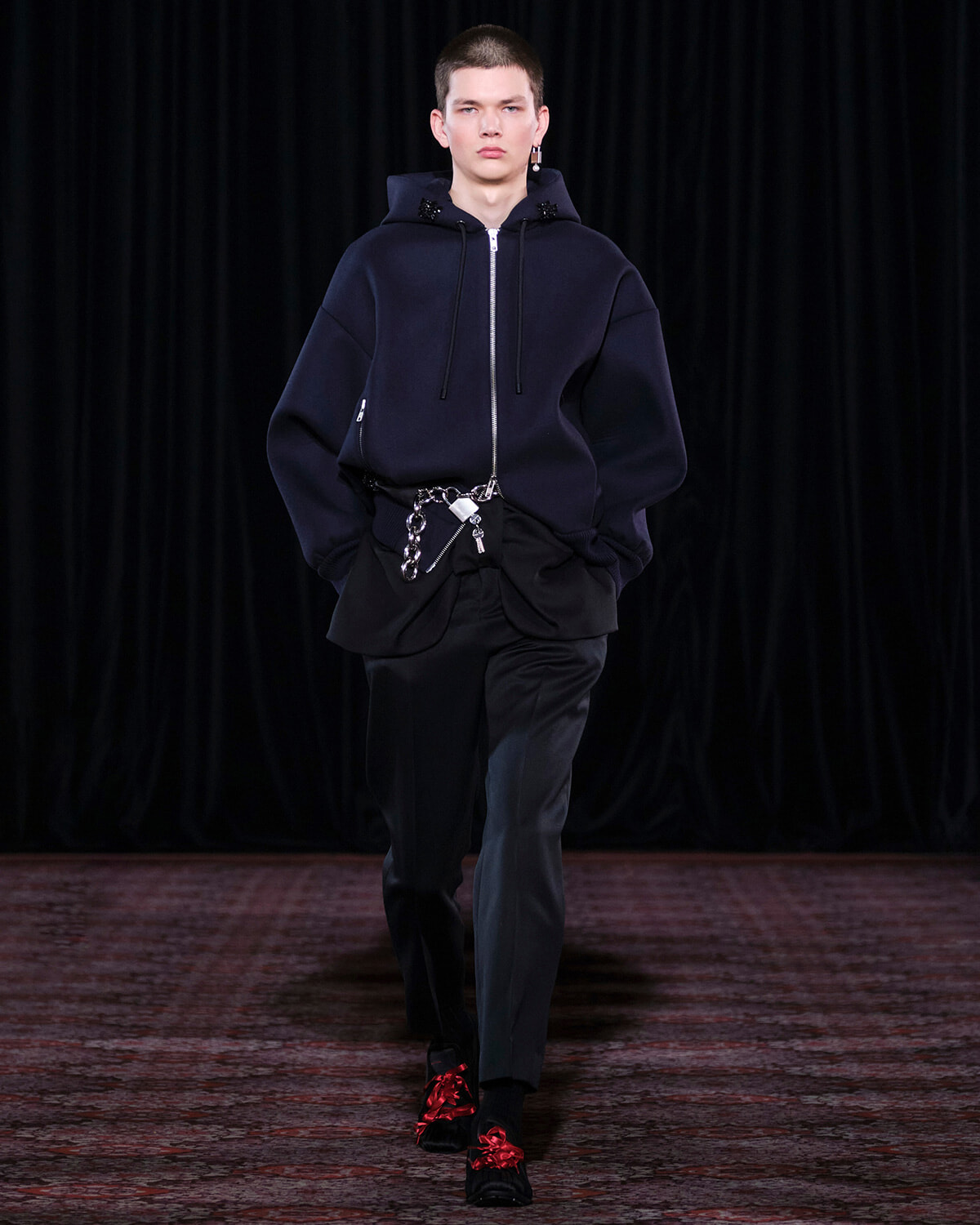 Male model walks the runway in an oversized navy hoodie with front zipper, black pants, a silver chain belt, and black shoes with red floral decorations.