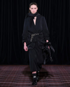 Model walking a dark, oversized black suit with a chain belt and a black scarf on a runway stage.