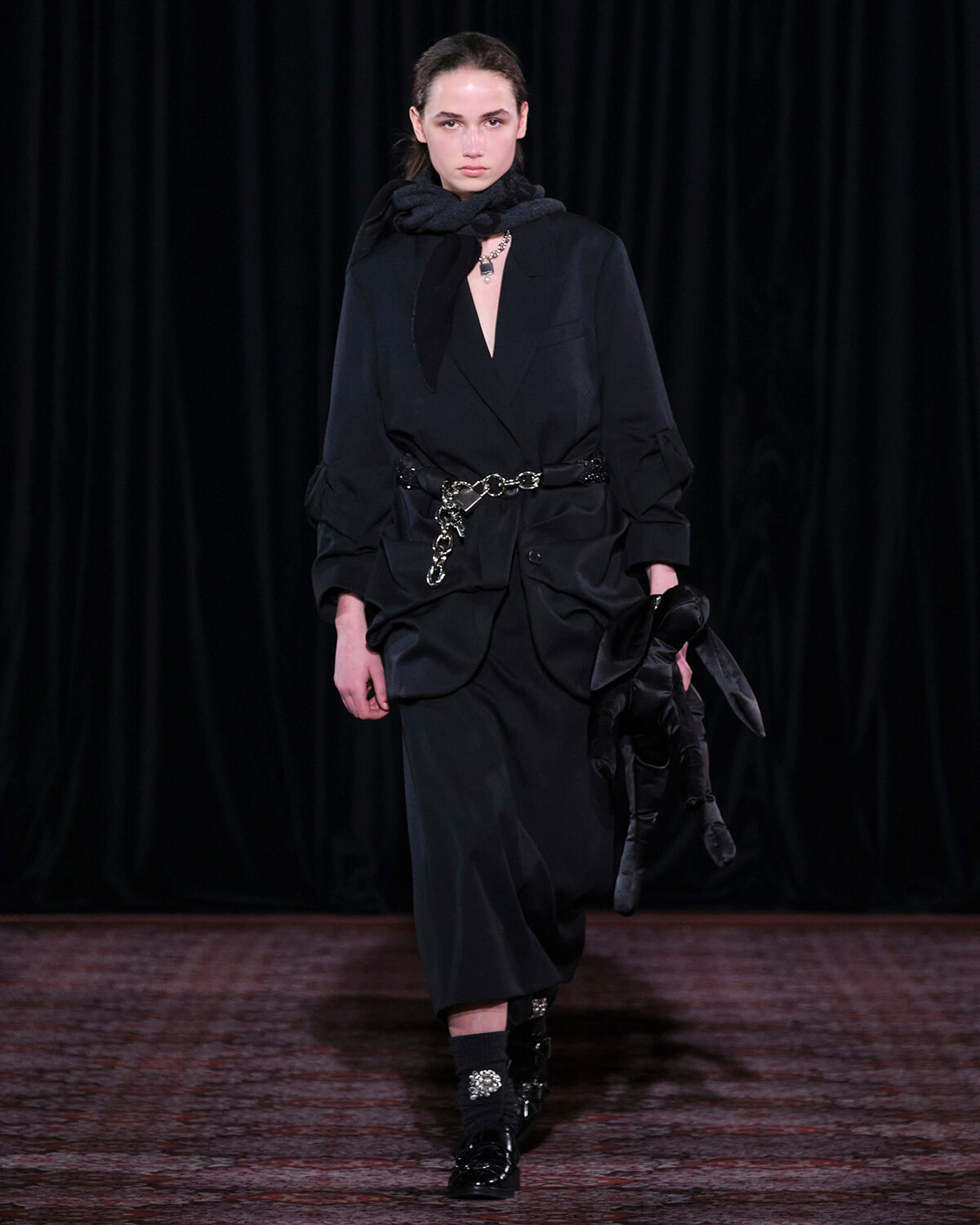 Model walking a dark, oversized black suit with a chain belt and a black scarf on a runway stage.
