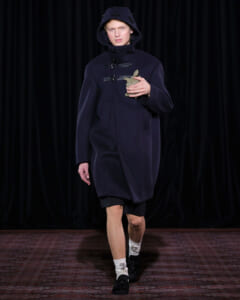 Model on a runway wearing a dark navy hooded coat with toggle fastenings, holding a small ceramic rabbit figurine in front of chest.