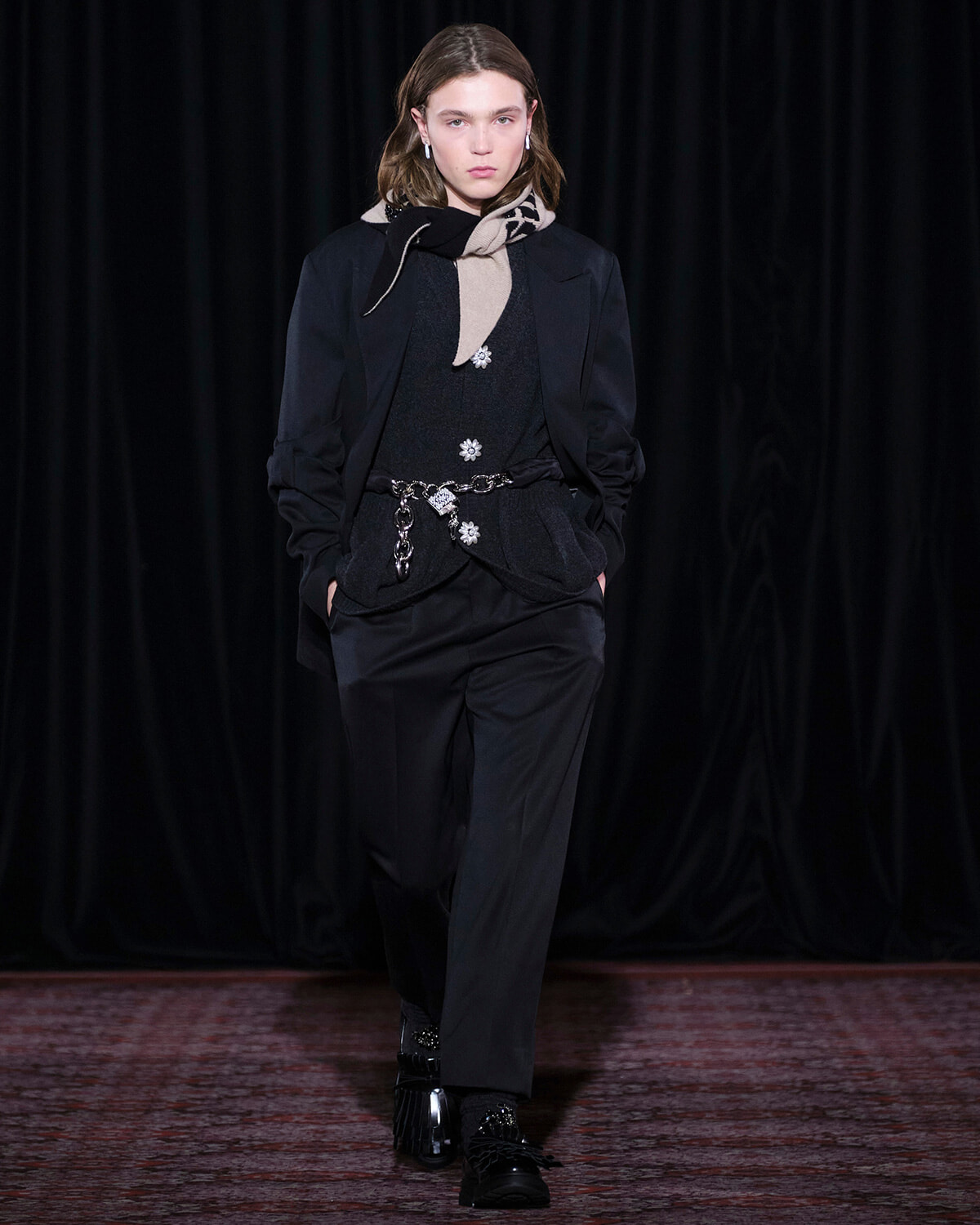 Model walking a fashion runway in a black suit with a scarf and silver belt accessories.