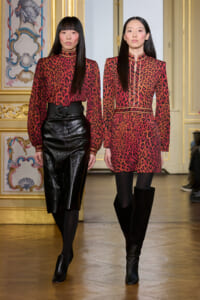 Two models in red leopard-print outfits walk a runway in a gilded room; one in a black leather skirt with a wide belt, the other in a matching dress with boots.