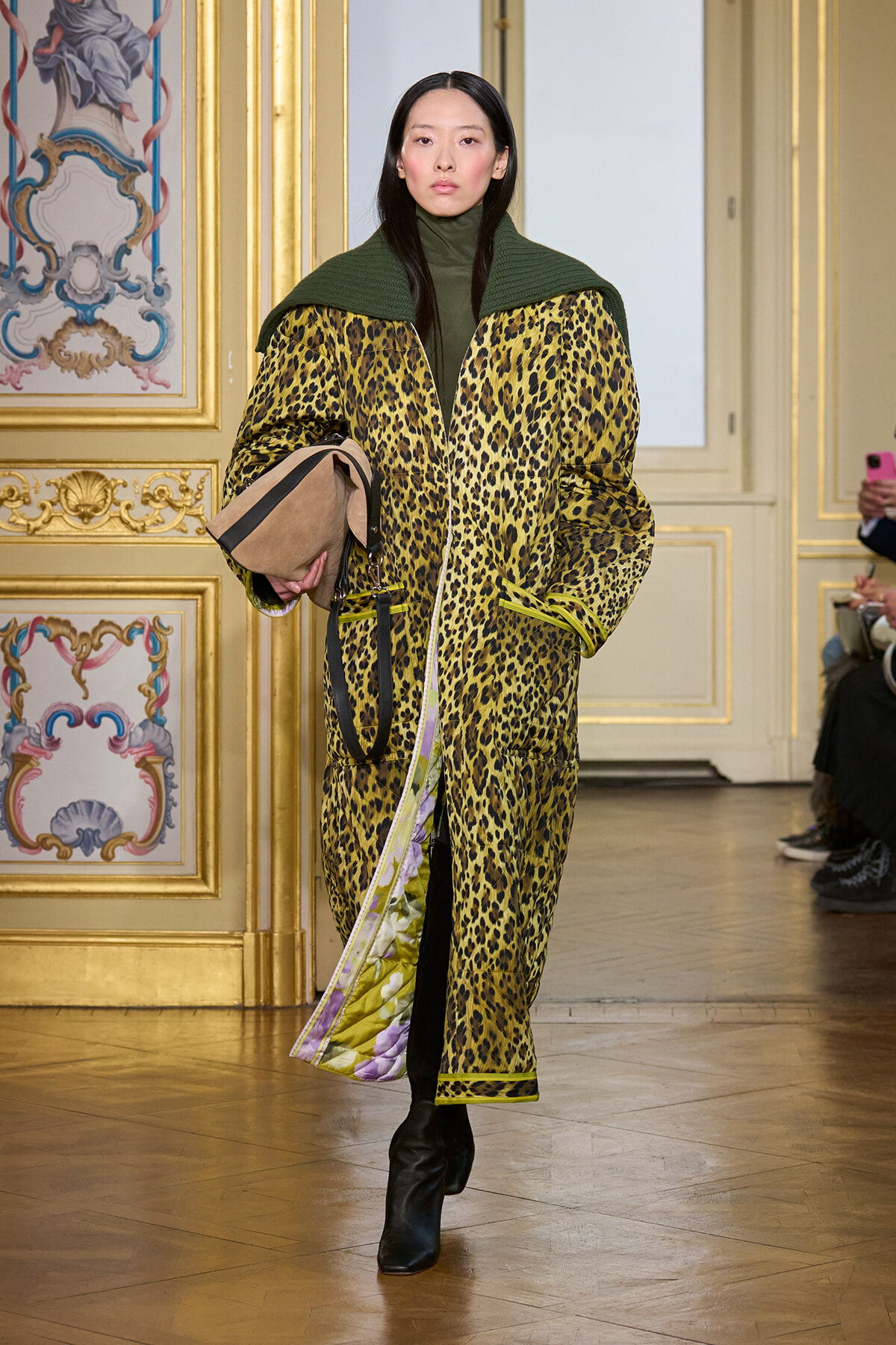 Model walks a fashion show in a bold leopard-print coat with a green cape, carrying a tan clutch.