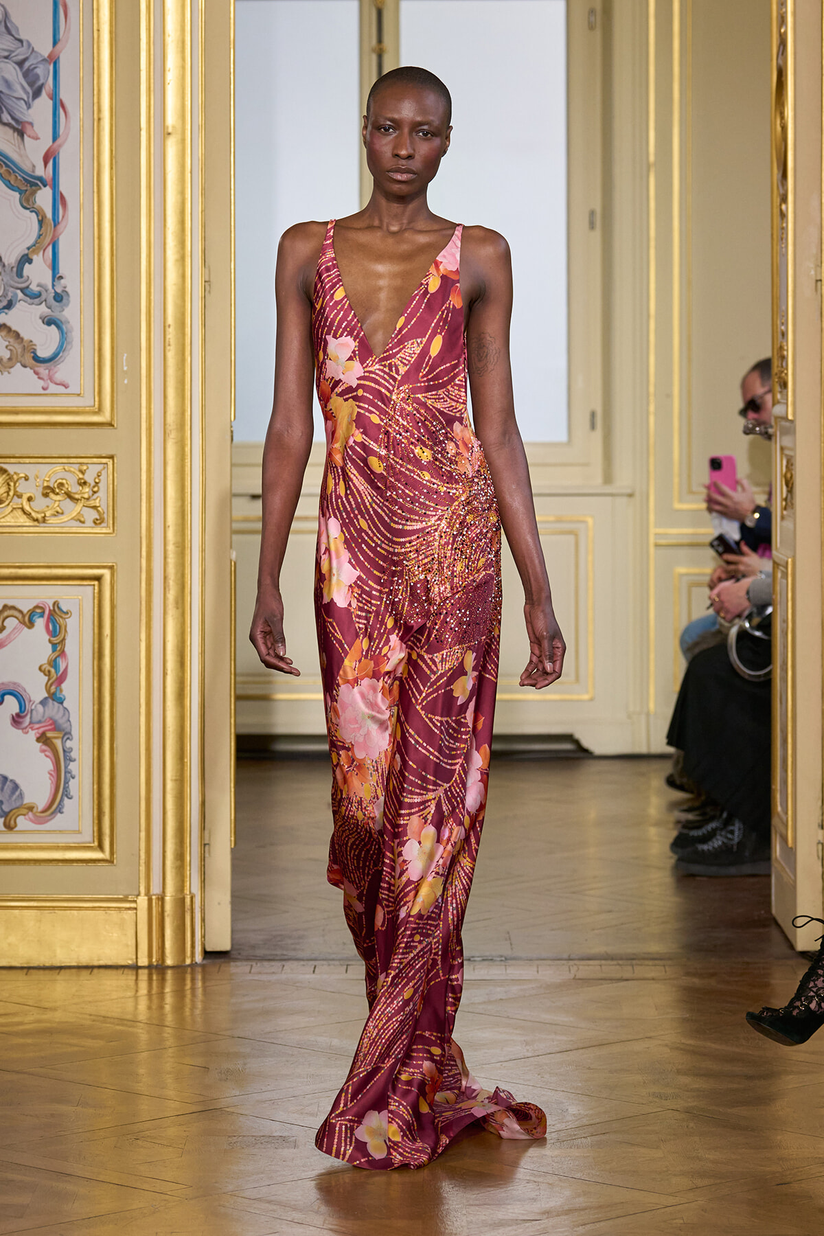 Model wearing a burgundy-pink floral gown with a deep V neckline walks down a gilded runway set in an ornate room