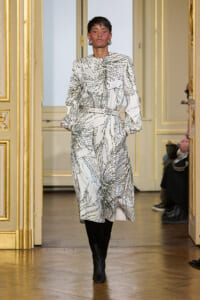 Model walking a runway in a white abstract-patterned midi dress with long sleeves and a cinched waist, paired with black knee-high boots, in a gilded room backdrop.