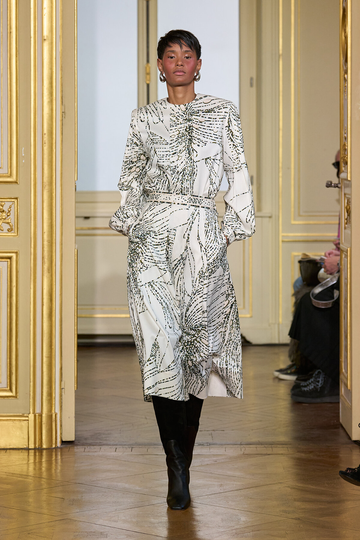 Model walking a runway in a white abstract-patterned midi dress with long sleeves and a cinched waist, paired with black knee-high boots, in a gilded room backdrop.
