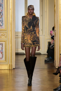 Runway model wearing a black and gold abstract print mini dress with matching headwrap and tall black boots, walking a gilded venue runway.