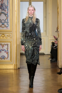Model walks a runway in a black and gold-patterned dress with a wide belt and black knee-high boots, in a gilded, ornate room.