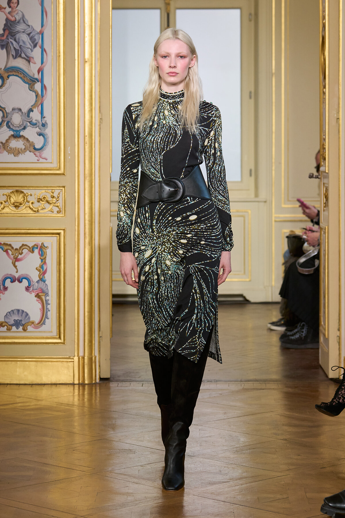 Model walks a runway in a black and gold-patterned dress with a wide belt and black knee-high boots, in a gilded, ornate room.
