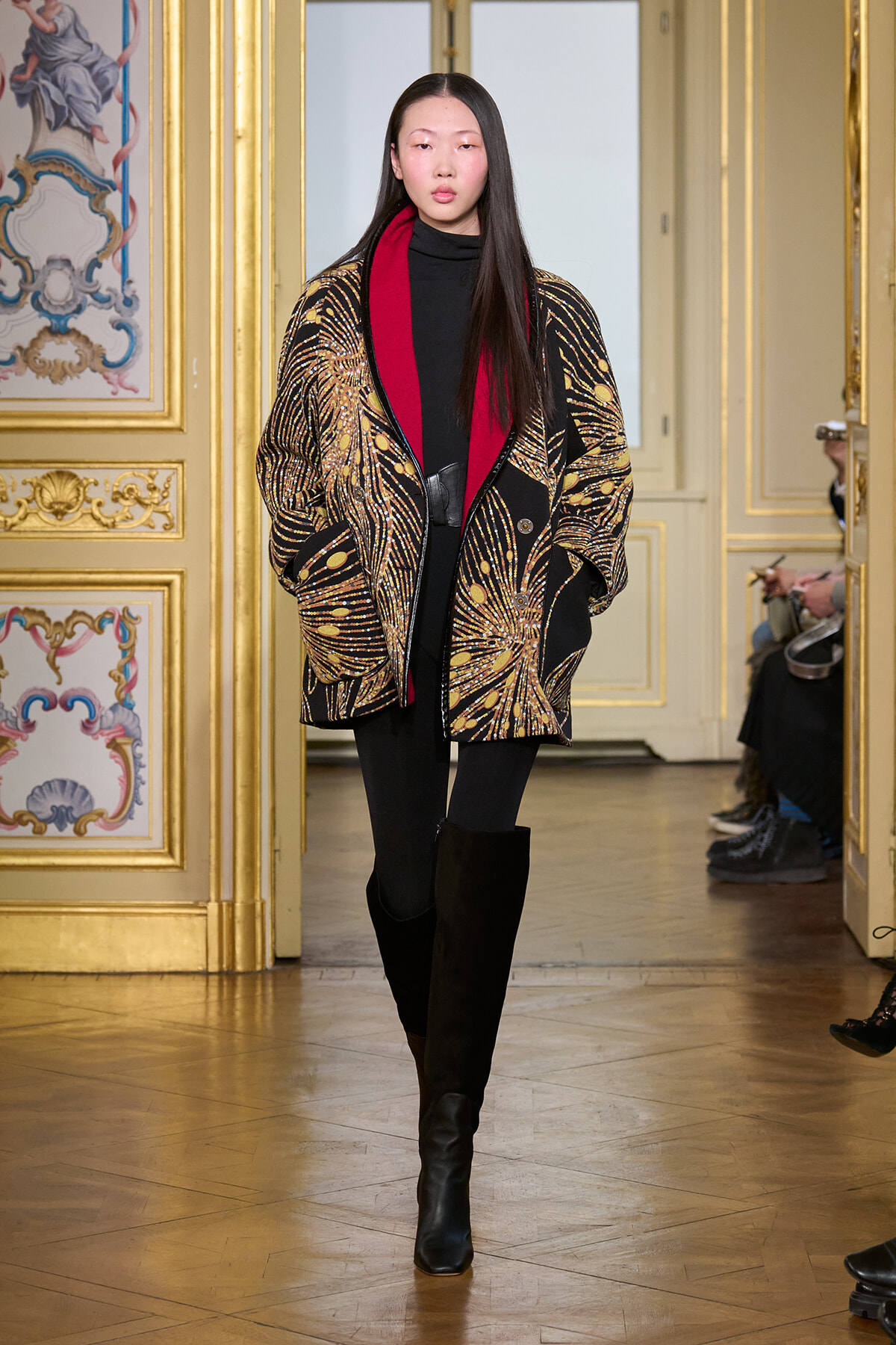 Model walks the runway in a black outfit with a gold-patterned cape and red lining, in a gilded, ornate room.