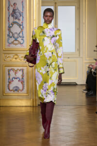Model walks in a gold-adorned room wearing a yellow-green floral turtleneck dress, burgundy knee-high boots, and holding a burgundy bag.