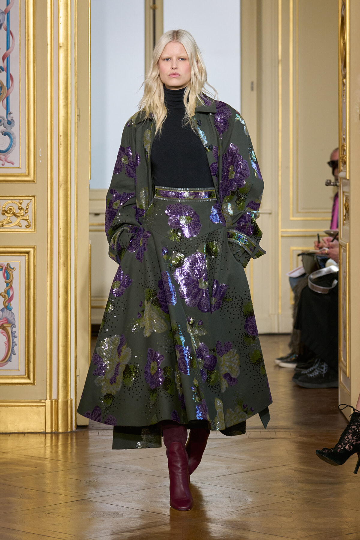 Model in a green floral sequined coat and matching skirt, black turtleneck, burgundy boots, walking a gilded fashion runway in a palatial room.