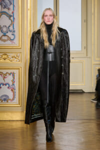Model on a runway wearing a long black textured coat over a black turtleneck and belt, in a gold-decorated room.