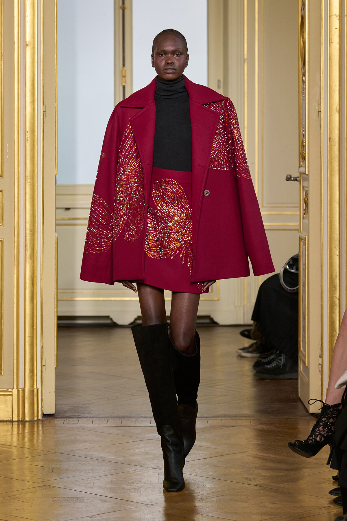Model walking a fashion show in a burgundy oversized coat with sequined patterns, paired with a black turtleneck and knee-high black boots in a gold-trimmed venue.