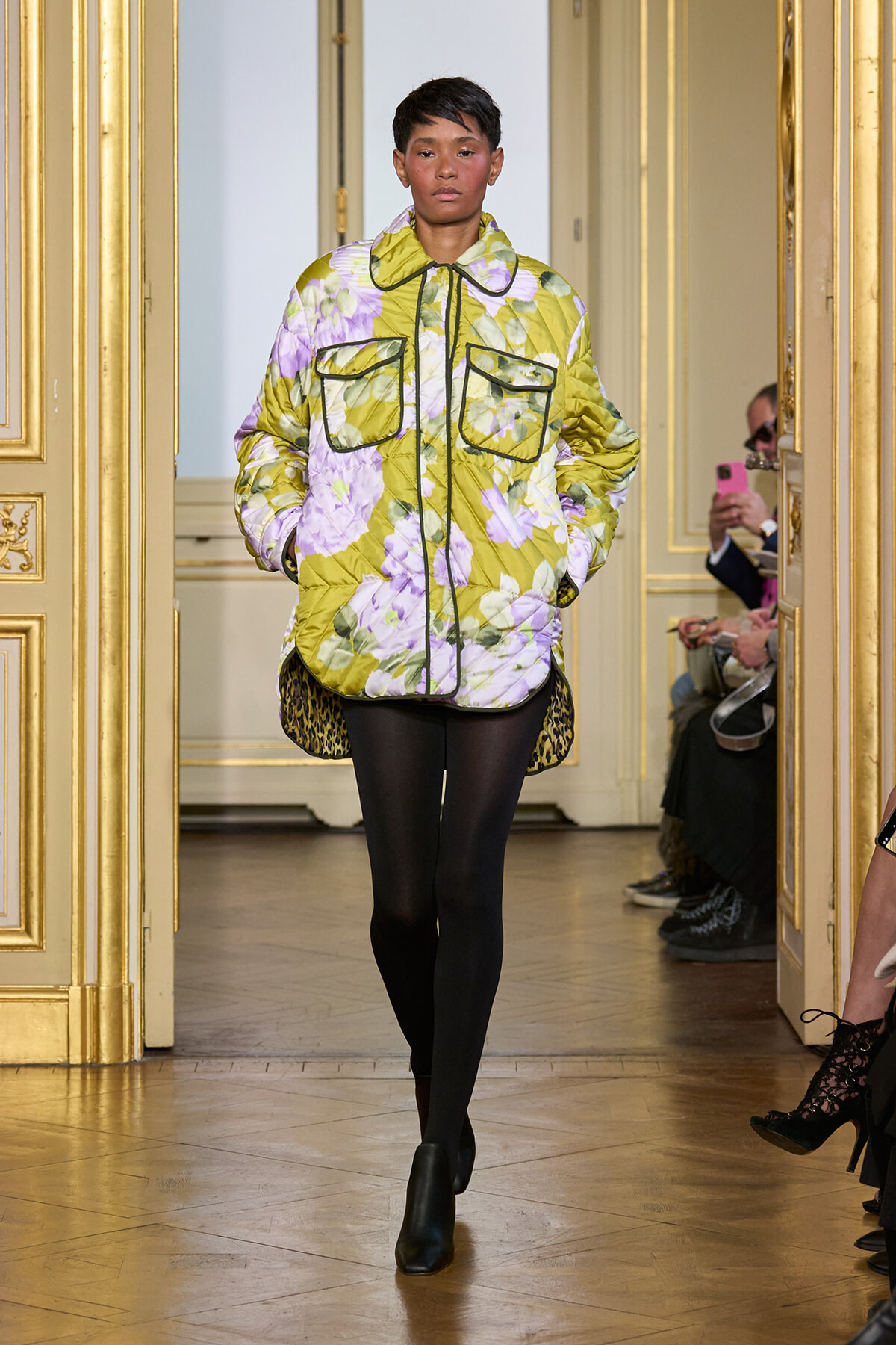 Model walks a runway in a oversized lime-green floral puffer jacket with large front pockets and black leggings.
