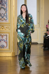 Model walking a runway in a long, loose dress with an abstract green, teal, and black geometric print in a gilded, ornate room.