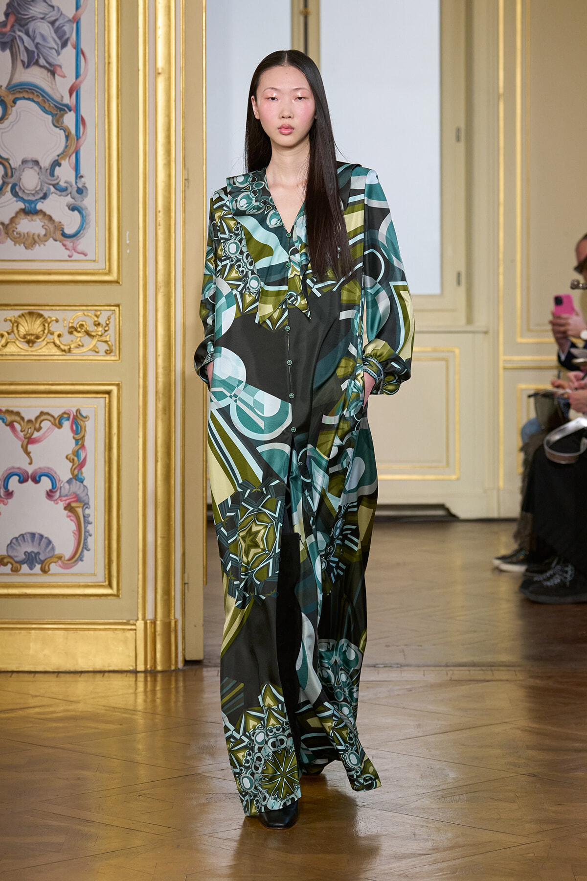 Model walking a runway in a long, loose dress with an abstract green, teal, and black geometric print in a gilded, ornate room.