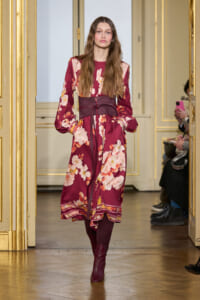Model walking a fashion runway wearing a burgundy floral midi dress with a wide burgundy belt and matching knee-high boots in a gilded venue.
