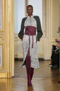 Model on a runway wearing a patterned long dress with green sleeves, a wide burgundy sash, and matching burgundy boots in a gilded room.
