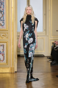 Model wearing a black floral jumpsuit with teal and white blossoms, cape-like shoulders, walking a fashion show down a gilded room runway.
