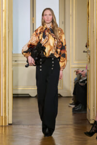 Model walks a runway in a bronze-toned patterned blouse with a neck bow and black high-waist trousers, carrying a black handbag.
