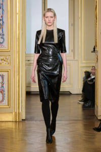 Model walking a knee-length black patent leather dress with a subtle floral pattern in a gilded, ornate room.