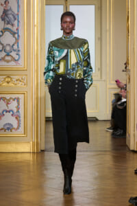 Model on a gilded catwalk wearing a green geometric blouse with high neck and black wide-leg trousers with silver buttons, in an ornate gold room.