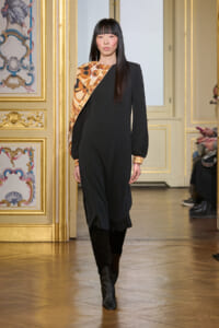 Model walks the runway in a black jumpsuit with an orange geometric sash in a gilded, ornate room setting.
