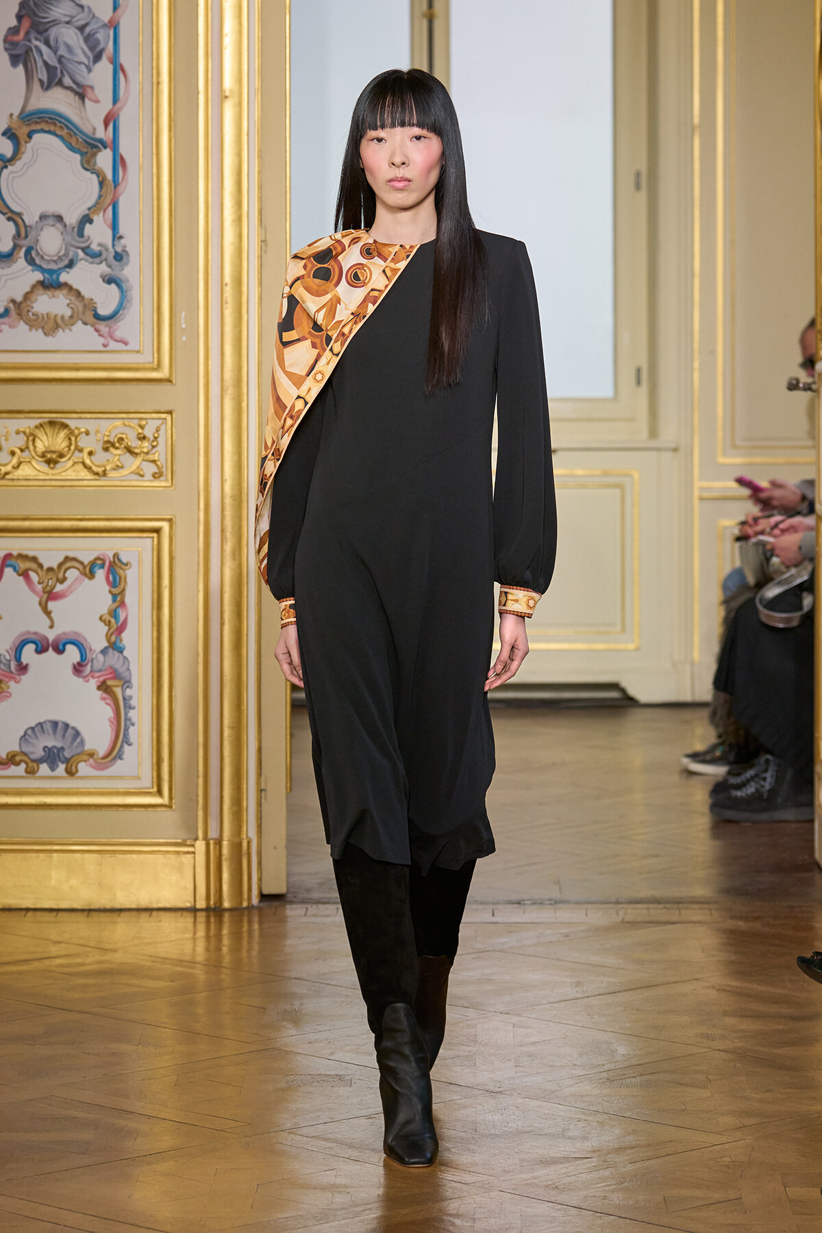Model walks the runway in a black jumpsuit with an orange geometric sash in a gilded, ornate room setting.