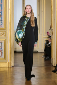 Model walking a runway in a black floor-length dress with a green geometric scarf draped over her left shoulder, in a gilded room.