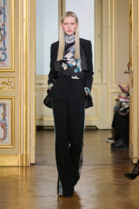 Model on a fashion runway in an ornate gold-trimmed room, wearing a black blazer over a floral turtleneck and black trousers with teal side piping.