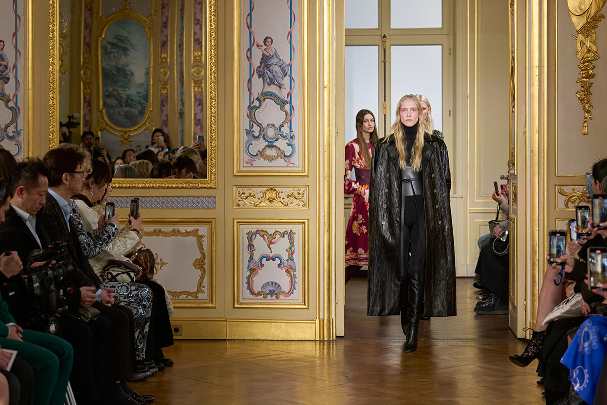 Model walks a catwalk in a black textured coat and knee-high boots inside a gold-decorated ballroom with seated audience photographing the scene.