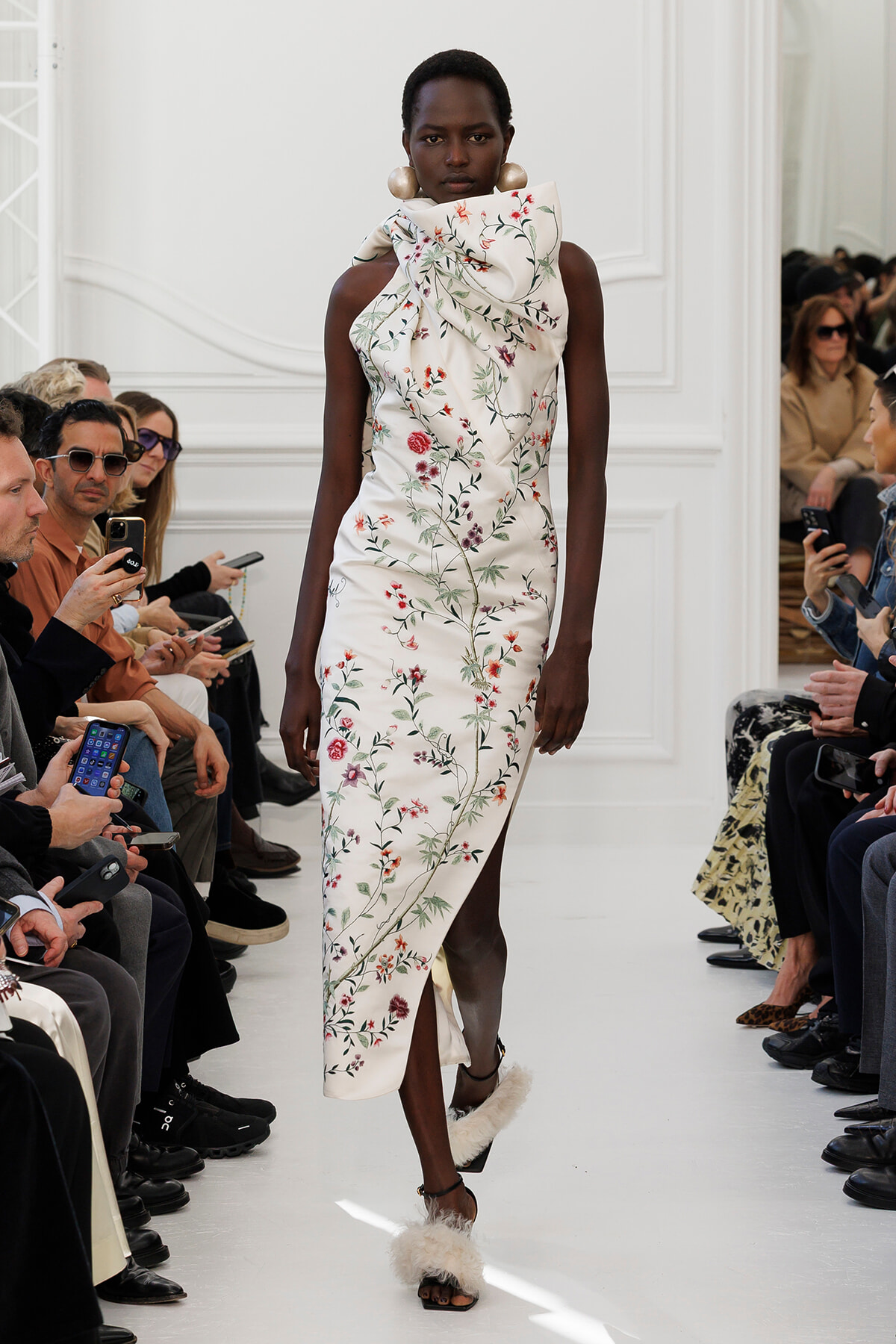 Black model walks a white runway in a sleeveless ivory floral gown with an oversized bow at the neck; audience sits along the sides.