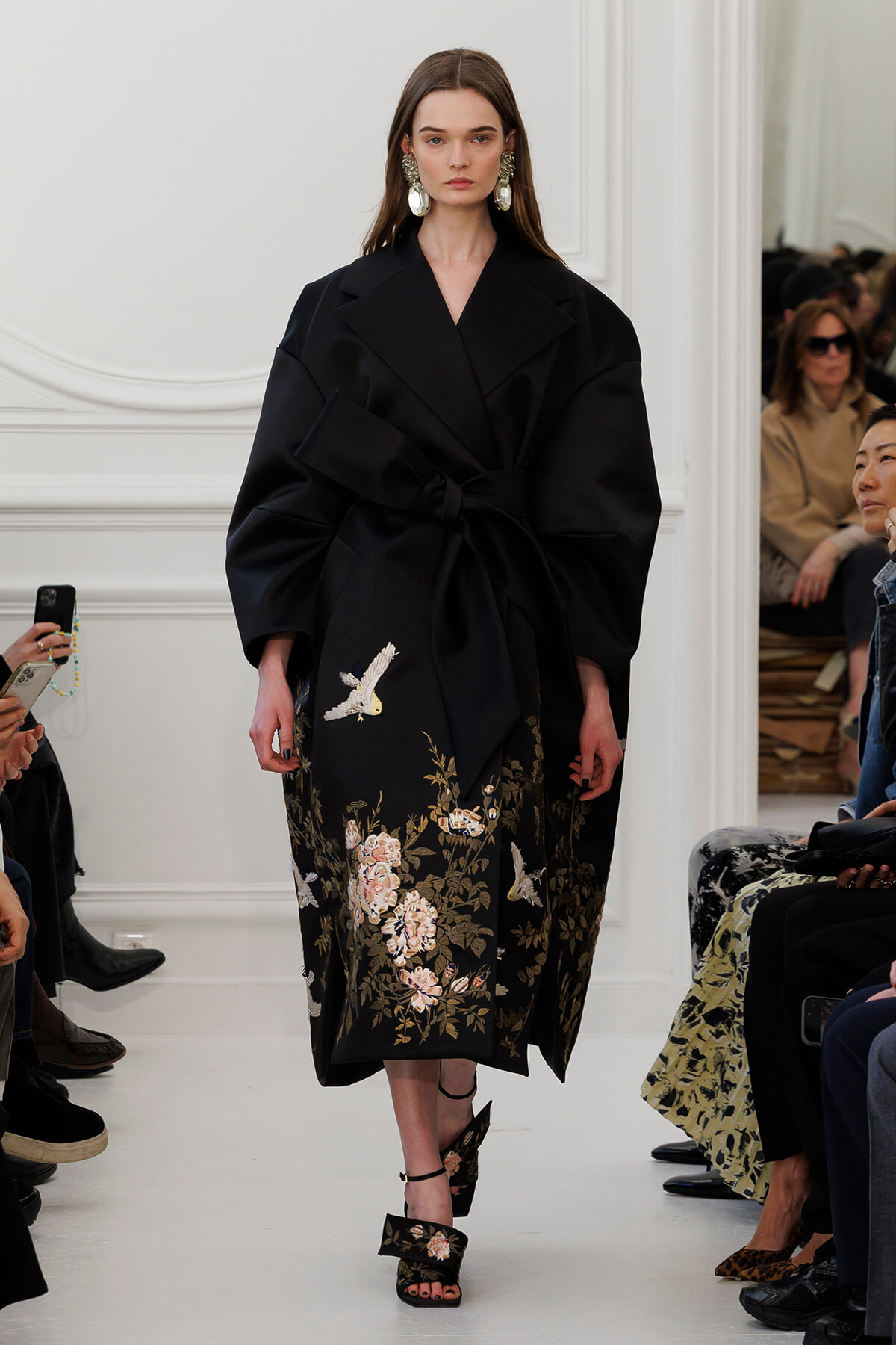 Model walking a runway in a black belted coat with large lapels and floral embroidery at the hem.
