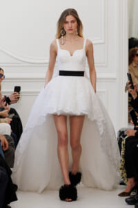 Runway model in a white lace mini dress with a black belt, and a long lace-train cape, accessorized with large dangling earrings.